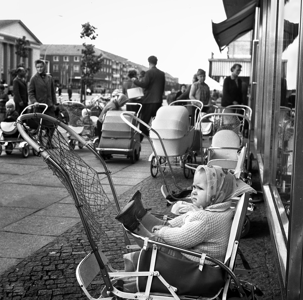 Kinderwagenparkplatz vor einer Verkaufseinrichtung in Eisenhüttenstadt 1962. Die Eltern mussten keine Sorge um die dort während des Einkaufs wartenden Kinder haben, die sich dort wohl fühlten. Das lässt sich an dem kleinen selbstbewussten Mädchen gut erkennen.
BArch Bild 183-A1120-010-001 008 Eisenhüttenstadt.jpg
